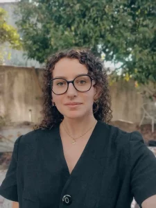 Portrait de Romane Bretbeil, chargée de communication, souriante avec ses cheveux bouclés et ses lunettes noires.