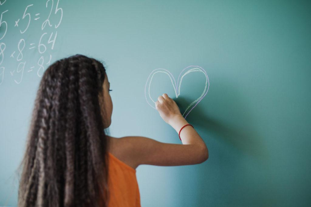 Écolière de dos dessinant un cœur à la craie sur un tableau, entre des tables de multiplication.